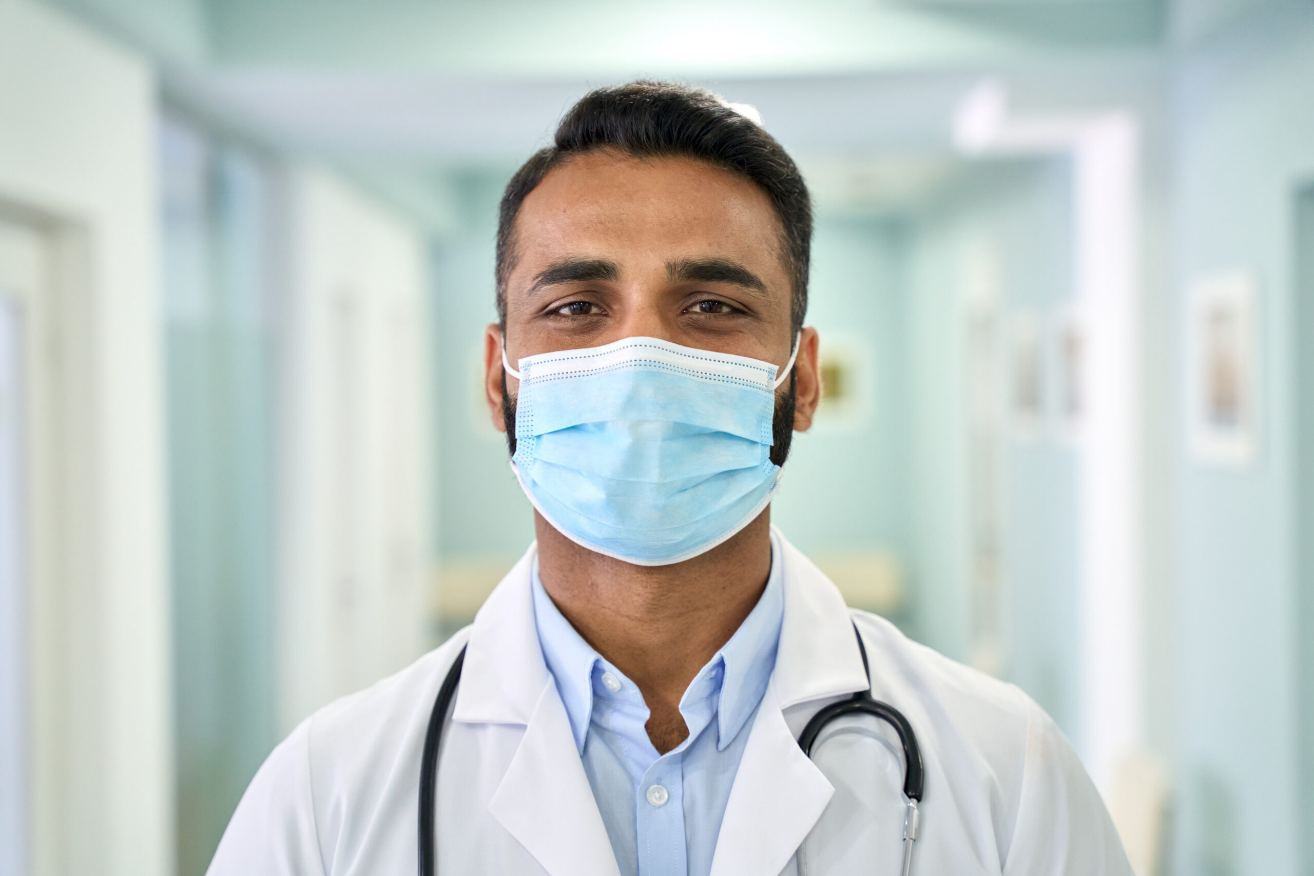 Male Indian doctor wearing medical coat and face mask looking at camera.
