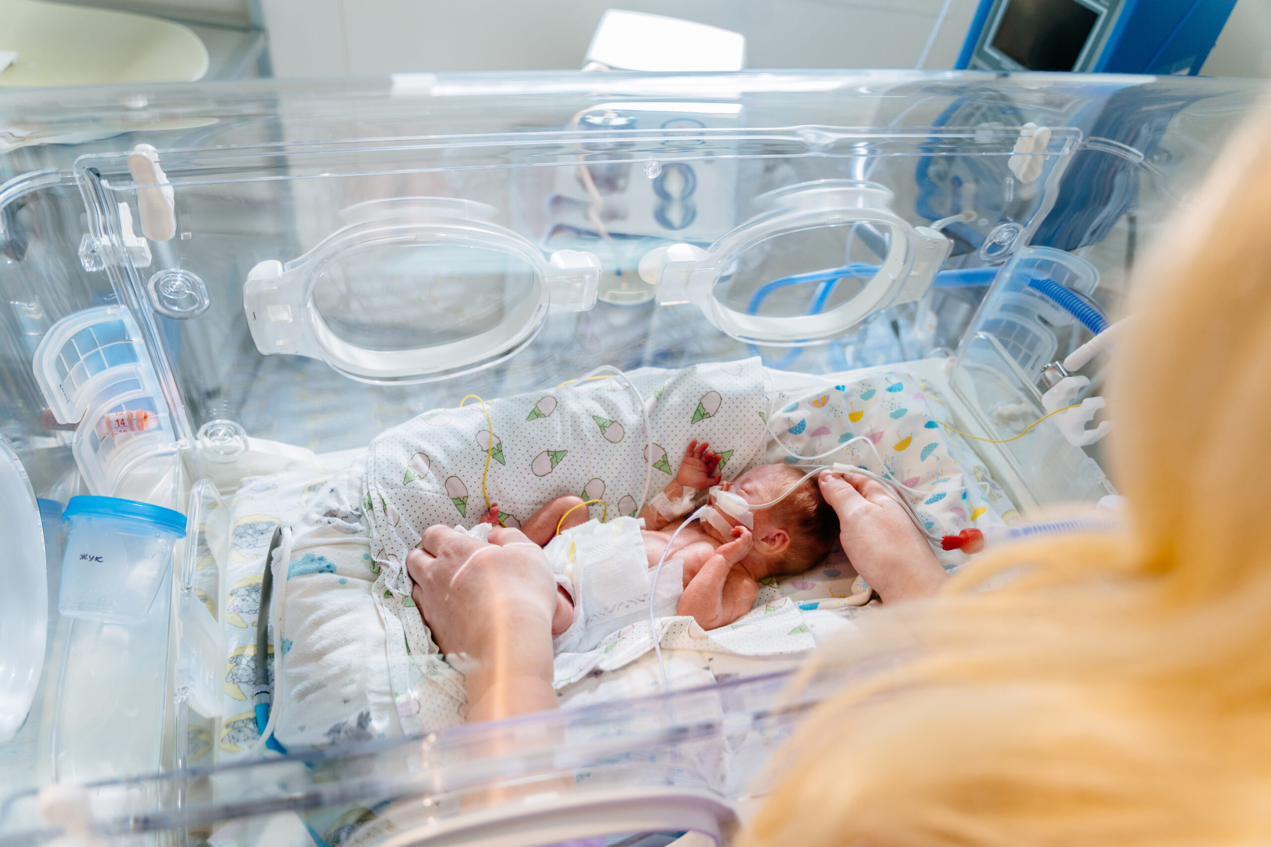 Unrecognizable mother’s hands holding new born baby born at 28 weeks gestation in intensive care unit in a medical incubator.